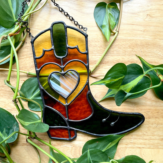 Green and orange stained glass cowboy boot on a wooden surface with plants 