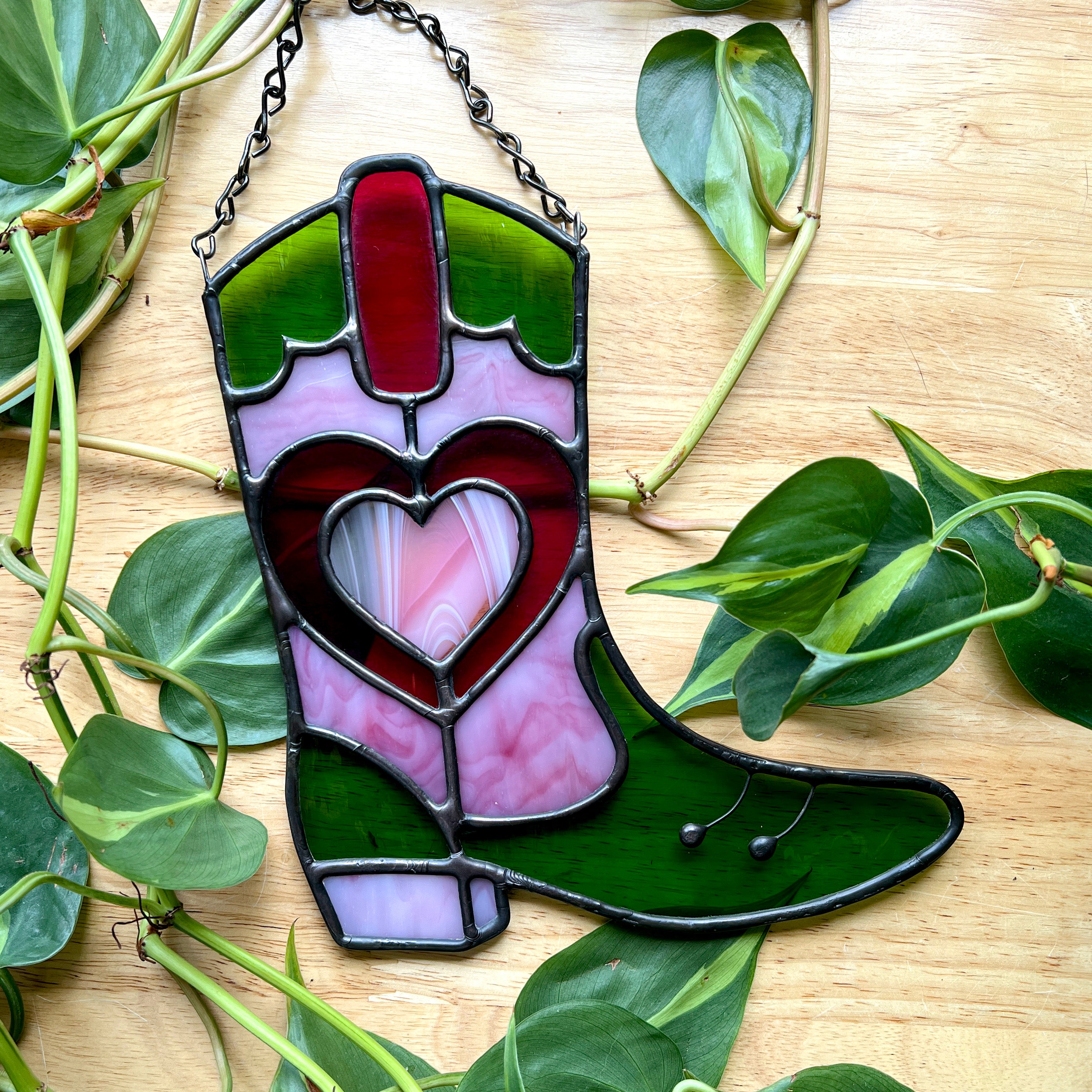 Pink and green stained glass cowboy boot on a wooden surface with plants 